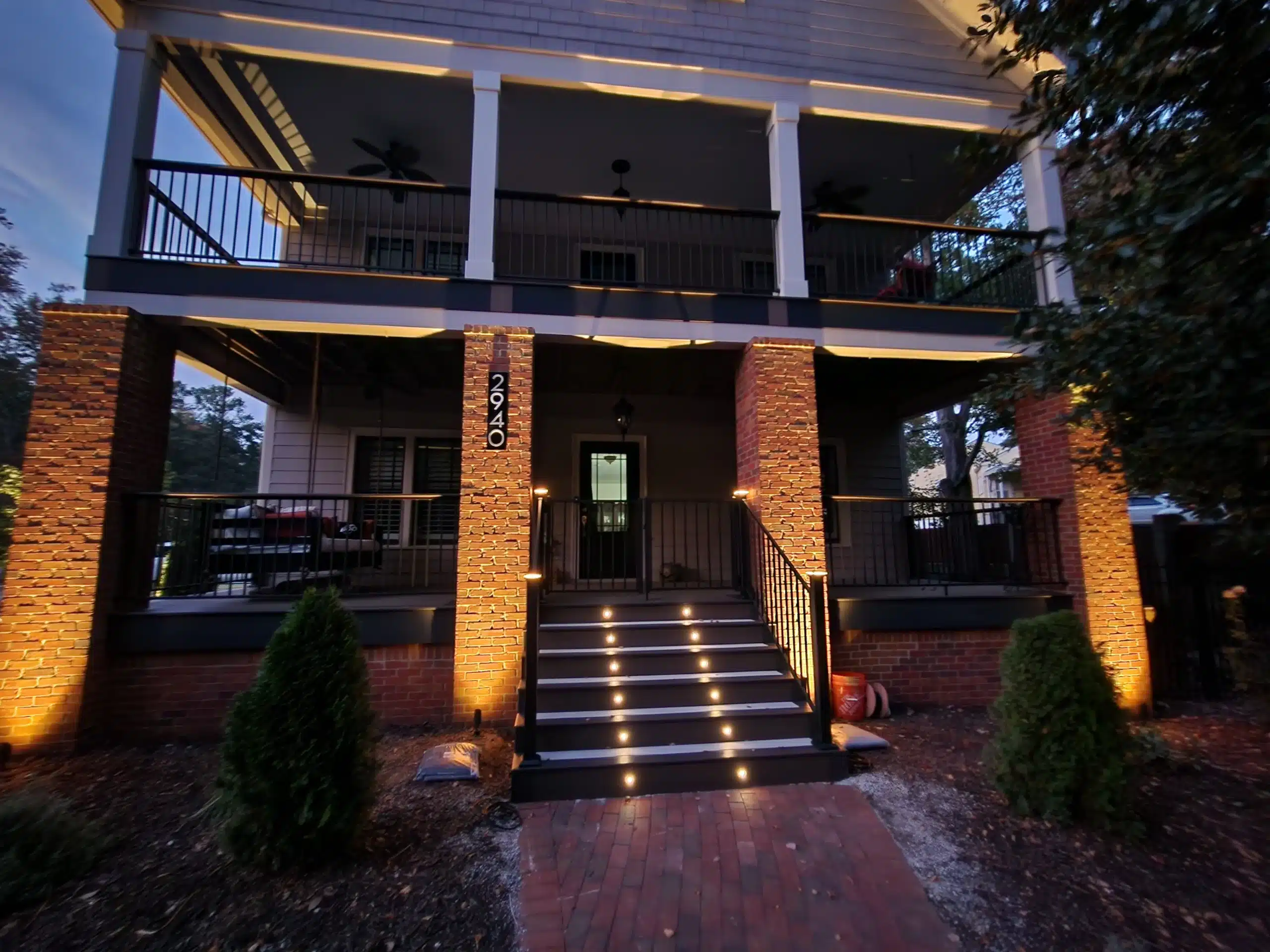 Two-level covered porch with brick columns and stair lighting, featuring black railings and recessed lighting.